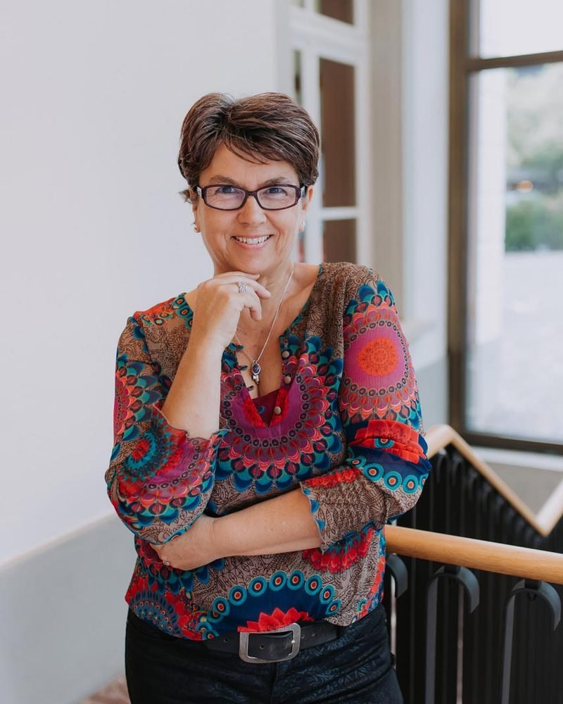 Claudia Doron in einem hellen Treppenhaus, lächelnd, mit Brille und farbig gemusterter Bluse.